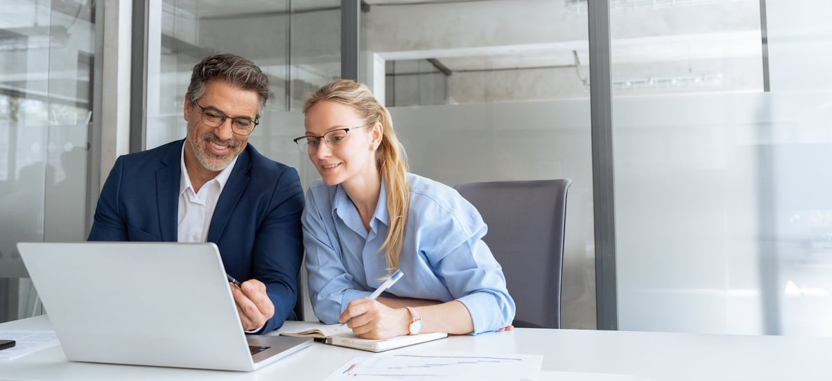 Mature mentor senior leader man showing young woman employee project on laptop in office.